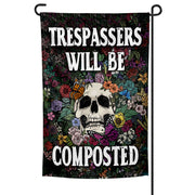 A garden flag with a skull and flowers design, featuring the text 'TRESPASSERS WILL BE COMPOSTED' on a black background.